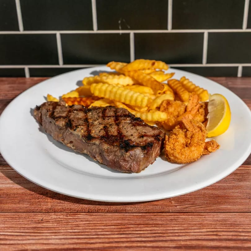 Grilled steak, crispy fried shrimp, crinkle-cut fries, and a lemon wedge on a white plate.