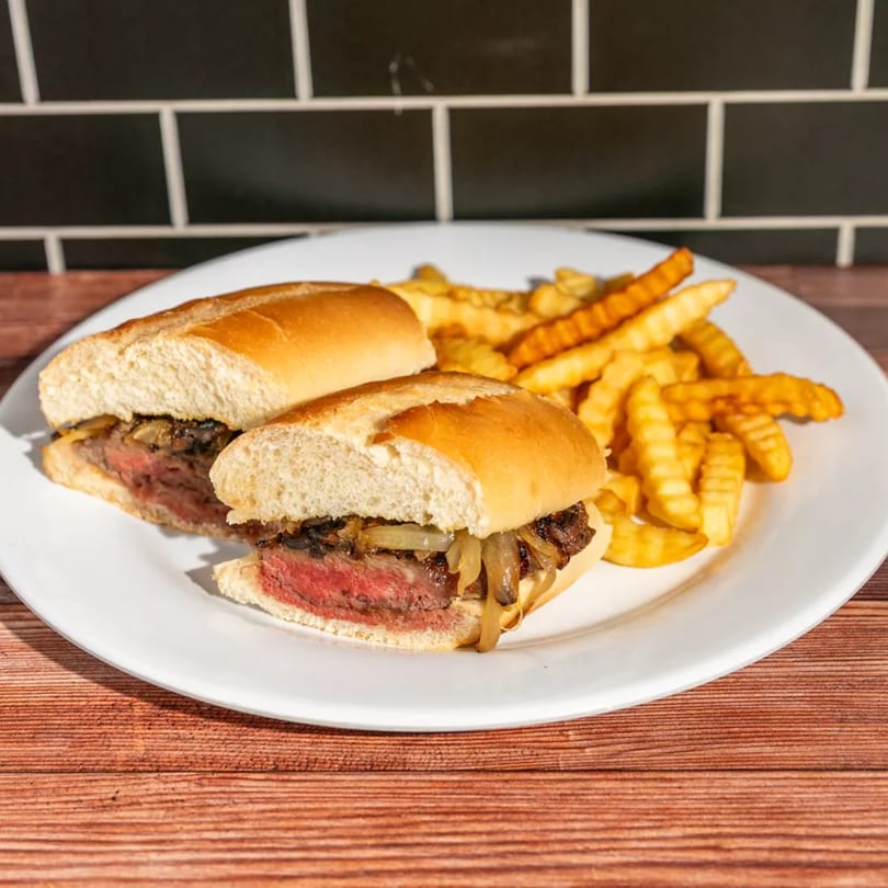 Steak sandwich with caramelized onions and a side of crinkle-cut fries on a white plate.